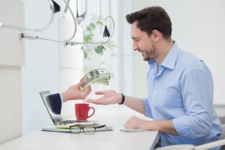 Happy businessman sitting in front of laptop computer while one hand holding  hundreds of dollar bills through laptop screen concept for internet e-commerce, paying and electronic banking.